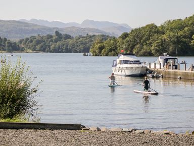 Two people paddleboarding on a lake with boats docked at a pier and hills in the background at Lake View Lodge in Lakeside 3 White Cross Bay Windermere