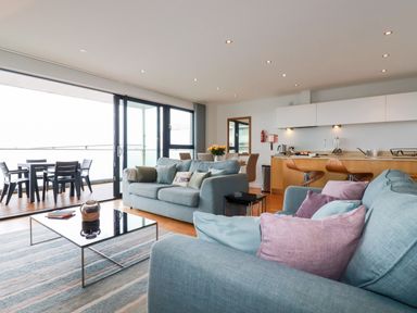 A living room with blue sofas and pink cushions a coffee table and a dining area with kitchen in the background at Fistral Pearl in Newquay