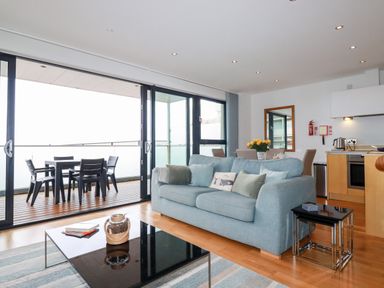 A living room with a blue sofa and coffee table next to a balcony with outdoor dining furniture at Fistral Pearl in Newquay