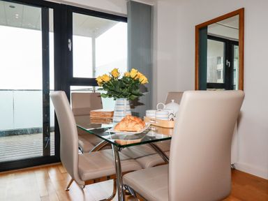 A dining area with a glass table, beige chairs, a vase with yellow flowers, and a plate with a croissant at Fistral Pearl in Newquay