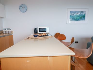 A kitchen island with pastries a microwave coffee machine and wooden chairs at Fistral Pearl in Newquay