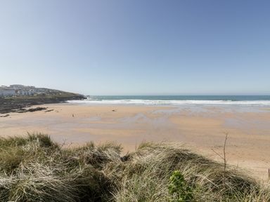 A sandy beach with waves and grass in the foreground and buildings on a hill to the left at Fistral Pearl in Newquay