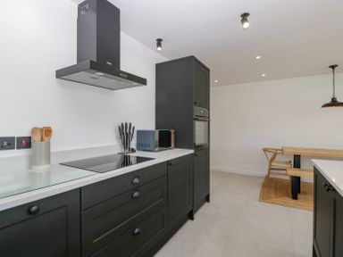 A kitchen with black cabinets white countertops a stovetop a microwave and a wooden dining table at Basswood Barn in Lime Tree Barn