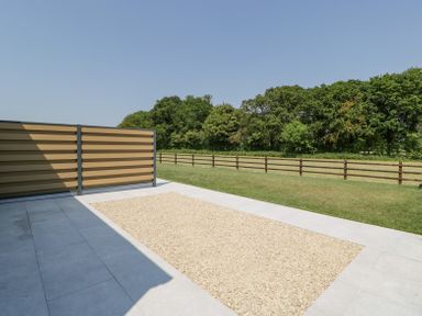 An outdoor patio area with a section of gravel and fencing overlooking a grassy field and trees at Basswood Barn in Lime Tree Barn