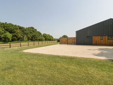 A building with wooden and metal siding next to a gravel area and grass field with a wooden fence and trees in the background at Basswood Barn in Lime Tree Barn