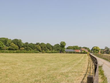 A grassy field next to a narrow road with a wooden fence and houses in the background at Basswood Barn in Lime Tree Barn