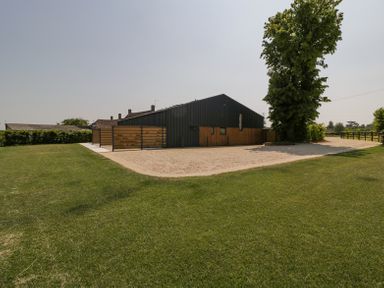 A large grassy area with a gravel path leading to a dark metal and wood building near a tall tree at Linden Barn in Wincanton