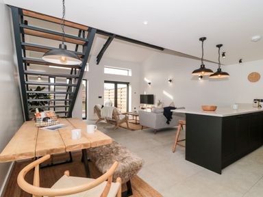 An open plan living area with a wooden dining table, staircase, grey sofa, armchairs, kitchen island with stools, and pendant lights at Linden Barn in Wincanton
