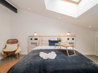 A bedroom with a bed with pillows and a blanket a wooden tray with a plant and a book a rattan chair with cushions and a window on the ceiling at Linden Barn in Wincanton