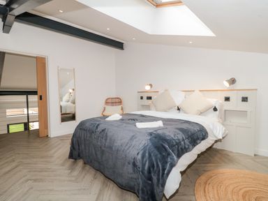 A bedroom with a double bed covered by a dark gray blanket two cushions on the bed a wicker chair a full-length mirror a round woven rug a skylight above the bed at Linden Barn in Wincanton