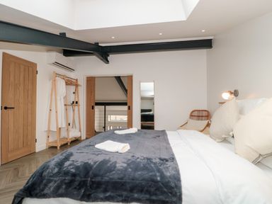 A bedroom with a large bed covered with a gray blanket and white pillows two wooden doors a wooden clothes rack with white robes a rattan chair and a full-length mirror at Linden Barn in Wincanton