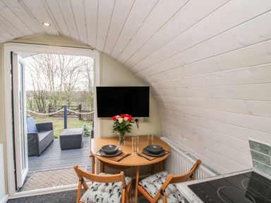 A small dining area with a table set for two and a TV on the wall next to an open door leading to an outdoor seating area at Cherry Pod 2 Hookagate near Meole Brace