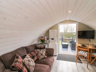 A room with a brown sofa with cushions a small dining table with two chairs a wall-mounted TV and a door leading outside at Cherry Pod 2 Hookagate near Meole Brace