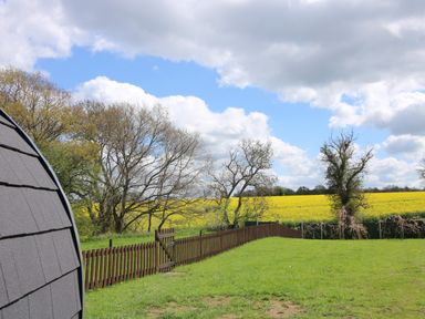 A fenced grassy garden area with trees and yellow fields under a partly cloudy sky at Cherry Pod 2 Hookagate near Meole Brace