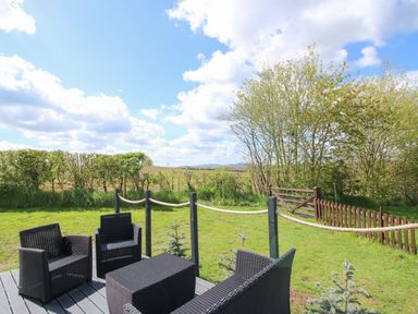 An outdoor patio with black wicker furniture and a rope fence overlooking a grassy area with trees and a wooden gate at Cherry Pod 2 Hookagate near Meole Brace