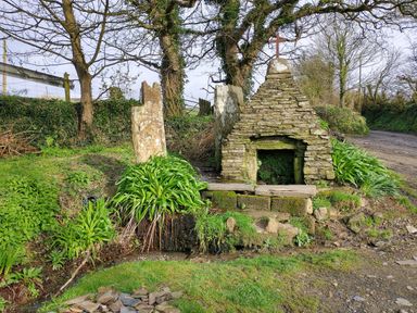 An outdoor stone structure with a cross on top surrounded by plants and trees near a dirt road at The Mill in Trethevy near Tintagel
