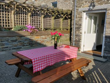 A wooden picnic table with a red and white checkered tablecloth with flowers and wine outside a stone building with open door at The Mill in Trethevy near Tintagel