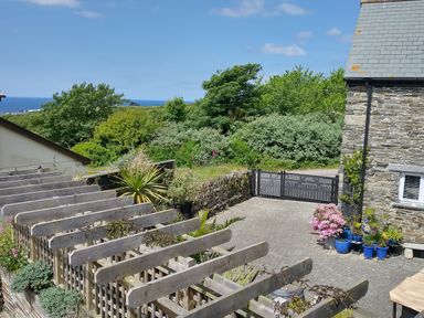 A paved driveway beside a stone house with blue pots and plants near a wooden pergola and green trees with an ocean view at The Mill in Trethevy near Tintagel