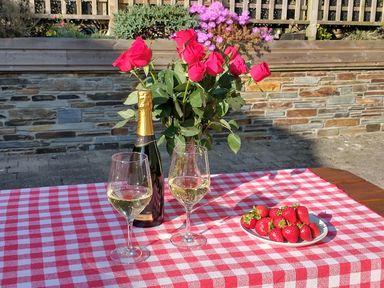A table with two glasses of white wine a bottle of champagne a vase of red roses and a plate of strawberries outdoors