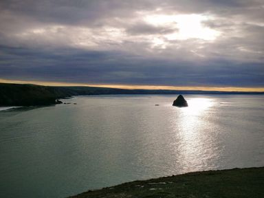 A coastal scene with cliffs to the left and a calm sea with a large rock in the water under a cloudy sky at The Mill in Trethevy near Tintagel