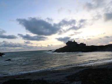 A silhouette of a house on a rocky peninsula by the ocean with waves and a cloudy sky at sunset at Porth Ewyn in Trearddur Bay