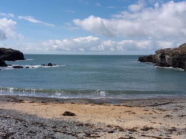 A coastal scene with a sandy and pebbly beach bordered by rocky cliffs and a calm sea at Porth Ewyn in Trearddur Bay