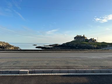 A coastal road with a rocky shoreline and a large stone house on a hill by the sea at Porth Ewyn in Trearddur Bay