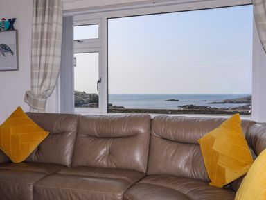 A living room with a brown leather sofa yellow cushions and a window showing a sea view at Porth Ewyn in Trearddur Bay