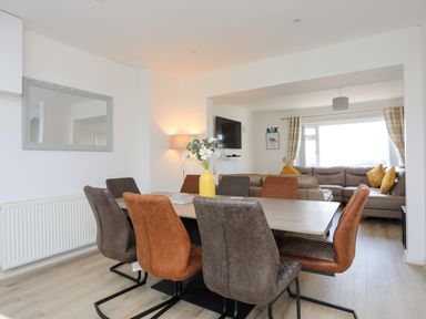 A dining area with a rectangular table and eight chairs next to a living room with a corner sofa and a wall-mounted TV at Porth Ewyn in Trearddur Bay