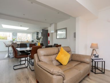 A living room with a brown leather sofa and yellow pillow next to a side table with a lamp and a dining area with chairs and a kitchen in the background at Porth Ewyn in Trearddur Bay