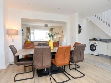 A dining area with a wooden table and six chairs next to a living room and kitchen at Porth Ewyn in Trearddur Bay