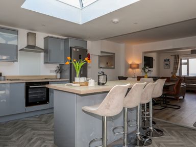 A kitchen with gray cabinets and an island with four white bar stools and orange flowers in a vase at Porth Ewyn in Trearddur Bay