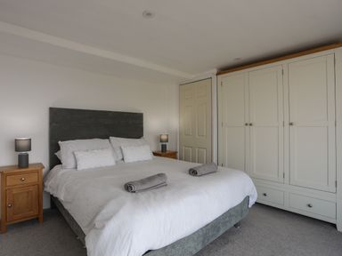 A bedroom with a double bed with white bedding grey headboard two bedside tables with lamps and a white wardrobe at Porth Ewyn in Trearddur Bay