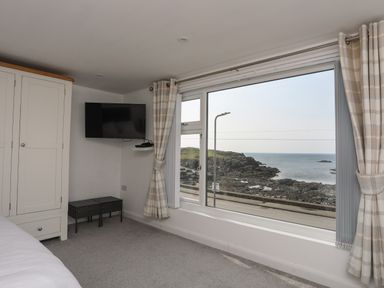 A bedroom corner with a wardrobe a wall-mounted television two stools large window with curtains and a sea view at Porth Ewyn in Trearddur Bay