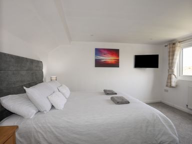 A bedroom with a large bed with pillows towels on it a wall-mounted tv a window with curtains and a framed picture on the wall at Porth Ewyn in Trearddur Bay