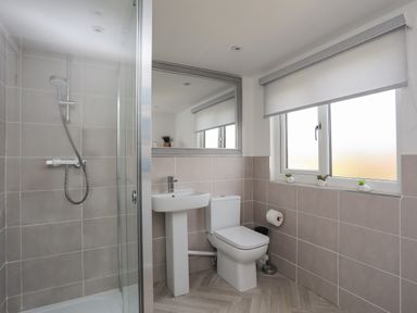 A bathroom with a shower, sink, toilet, window with blinds and small plants on the sill at Porth Ewyn in Trearddur Bay