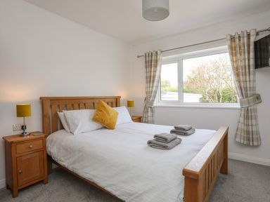 A bedroom with a wooden bed, bedside tables with lamps, folded towels on the bed, and a window with curtains at Porth Ewyn in Trearddur Bay