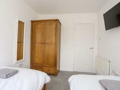 A bedroom with two beds with gray towels on them a wooden wardrobe a wall mirror a white door a wall-mounted TV and a radiator at Porth Ewyn in Trearddur Bay