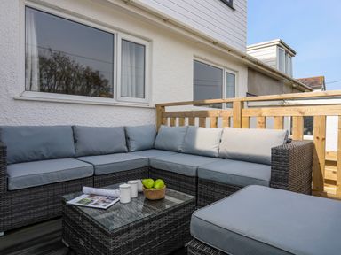 An outdoor seating area with a gray cushioned wicker corner sofa and a matching table with mugs and fruit basket at Porth Ewyn in Trearddur Bay