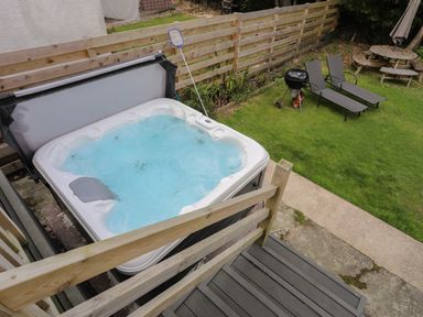An outdoor area with a hot tub near wooden railing a barbecue grill two lounge chairs and a picnic table on a grassy lawn at Porth Ewyn in Trearddur Bay