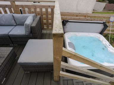 An outdoor seating area with grey cushioned wicker furniture and a wooden railing next to a hot tub with bubbling water at Porth Ewyn in Trearddur Bay