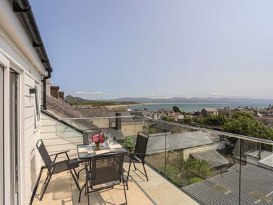 A balcony with a table and four chairs with flowers and glasses overlooking a town and the sea at Golygfa'r Gastell Castle View in Criccieth