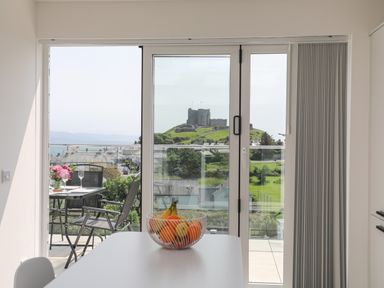 A dining area with a bowl of fruit on the table and a balcony with chairs and flowers overlooking a castle at Golygfa'r Gastell Castle View in Criccieth