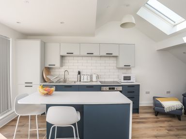A modern kitchen with blue and white cabinets a countertop with stools and a fruit bowl at Golygfa'r Gastell Castle View in Criccieth