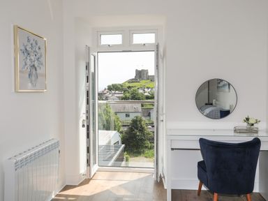 A room with an open glass door showing a castle outside a white desk with a round mirror and a blue chair at Golygfa'r Gastell Castle View in Criccieth