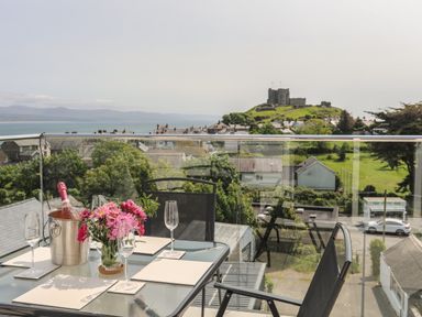A balcony with a glass table set with flowers glasses and a bottle of wine overlooking a castle on a hill at Golygfa'r Gastell Castle View in Criccieth