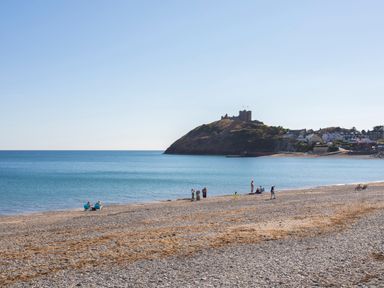 A pebble beach with people near the shoreline and a castle on a hill in the background at Golygfa'r Gastell Castle View in Criccieth