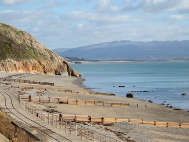 A coastal path with railings next to a rocky beach and calm sea with mountains in the background at Golygfa'r Gastell Castle View in Criccieth