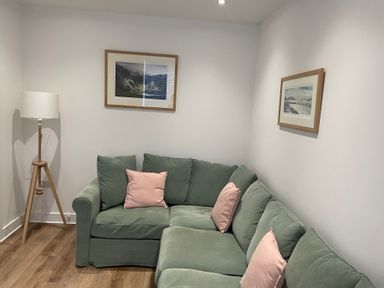 A corner of a room with a green sectional sofa with pink cushions two framed pictures on walls and a floor lamp with a wooden tripod base at Golygfa'r Gastell Castle View in Criccieth