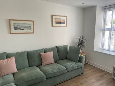 A green sofa with pink cushions next to a window with blinds and two framed pictures on the wall at Golygfa'r Gastell / Castle View in Criccieth